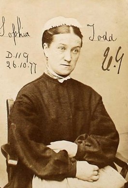 A woman in a black dress with white cap sits with her arms folded in her lap