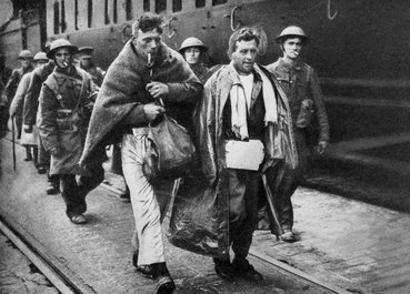 Two men covered in blankets, one smoking a pipe, walk ahead of helmeted soldiers in uniform.